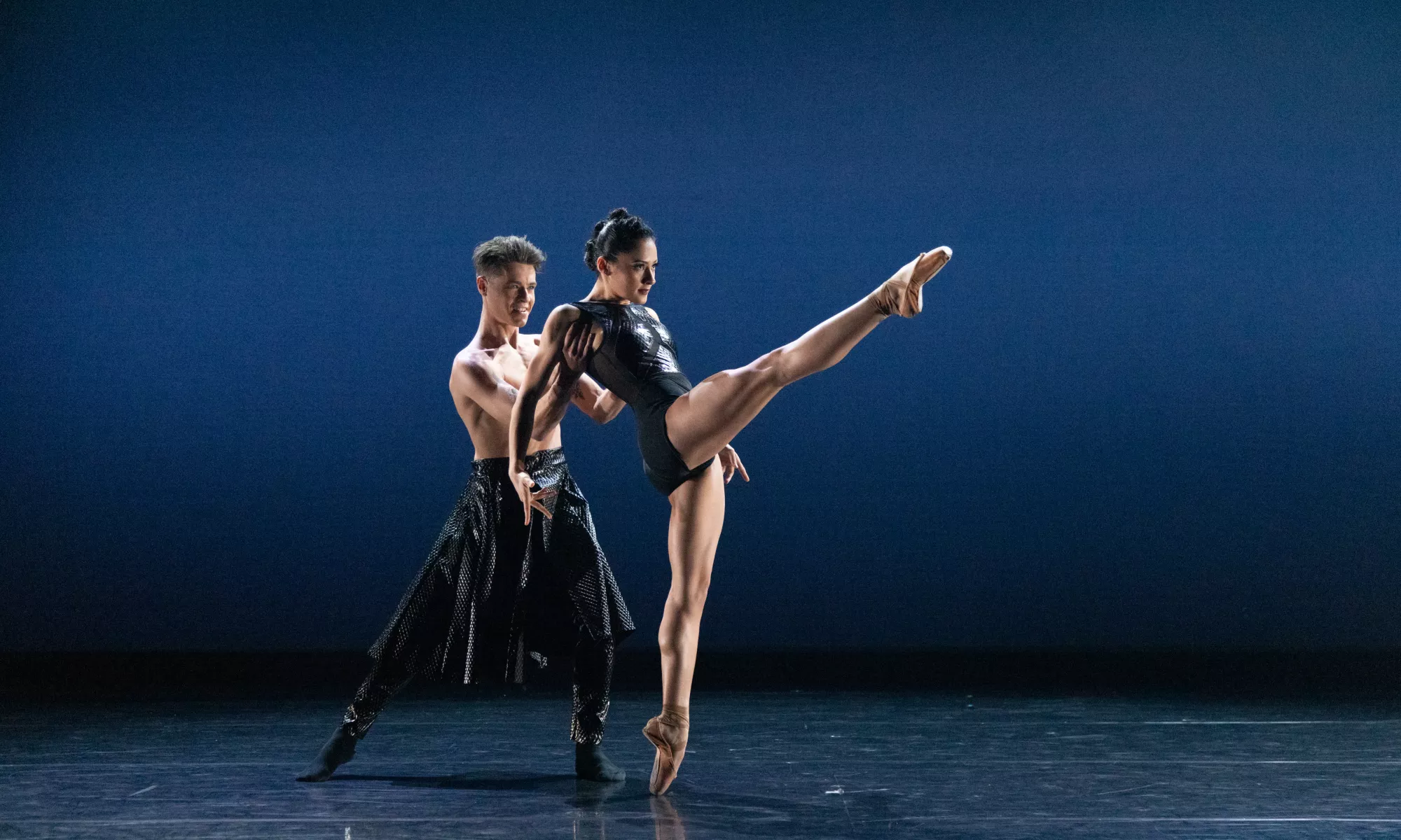 Two dancers performing. The ballerina has her leg extended while slightly leaning back onto the arms of the male dancer.