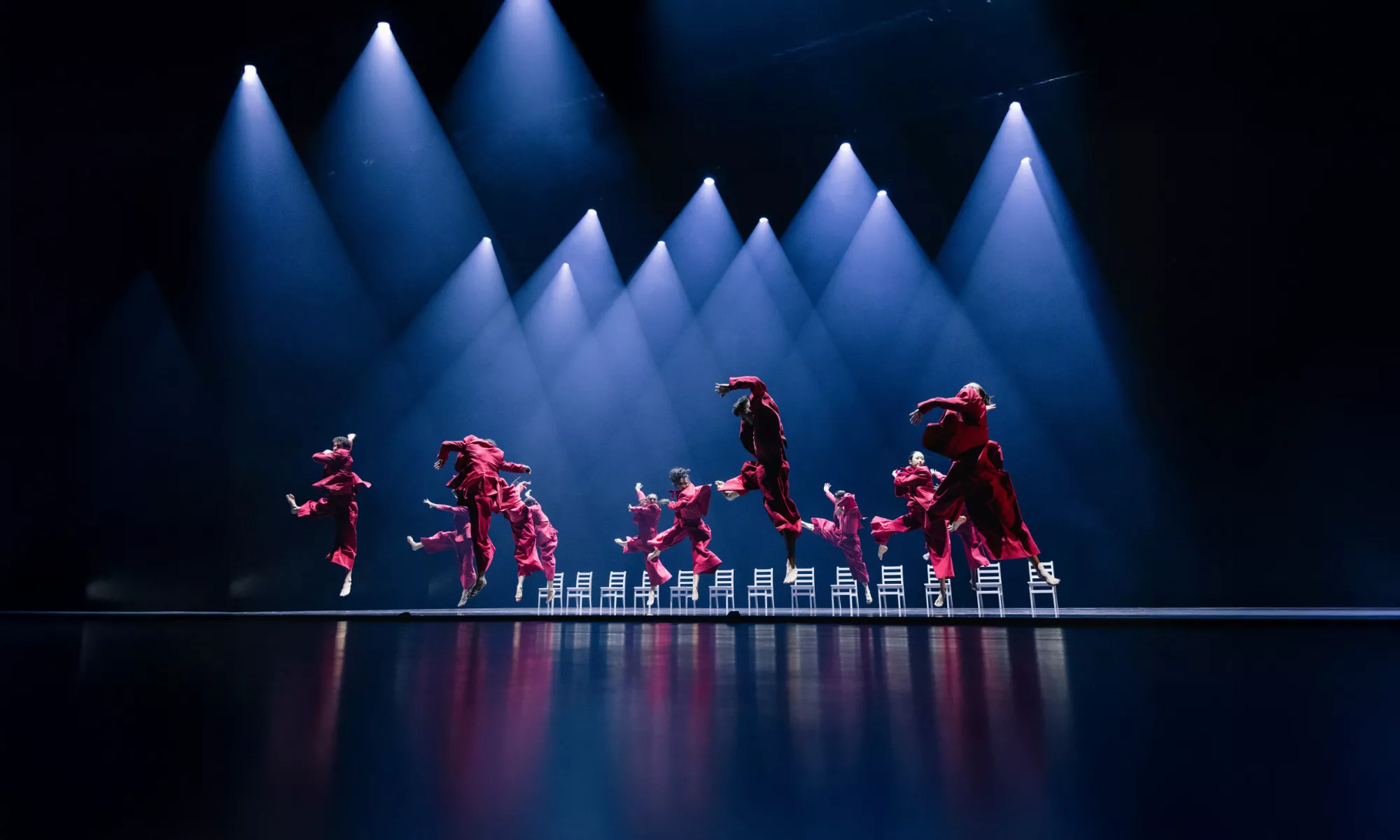 Dancers wear red suits and jump in the air. There is a line of white chairs in the background.