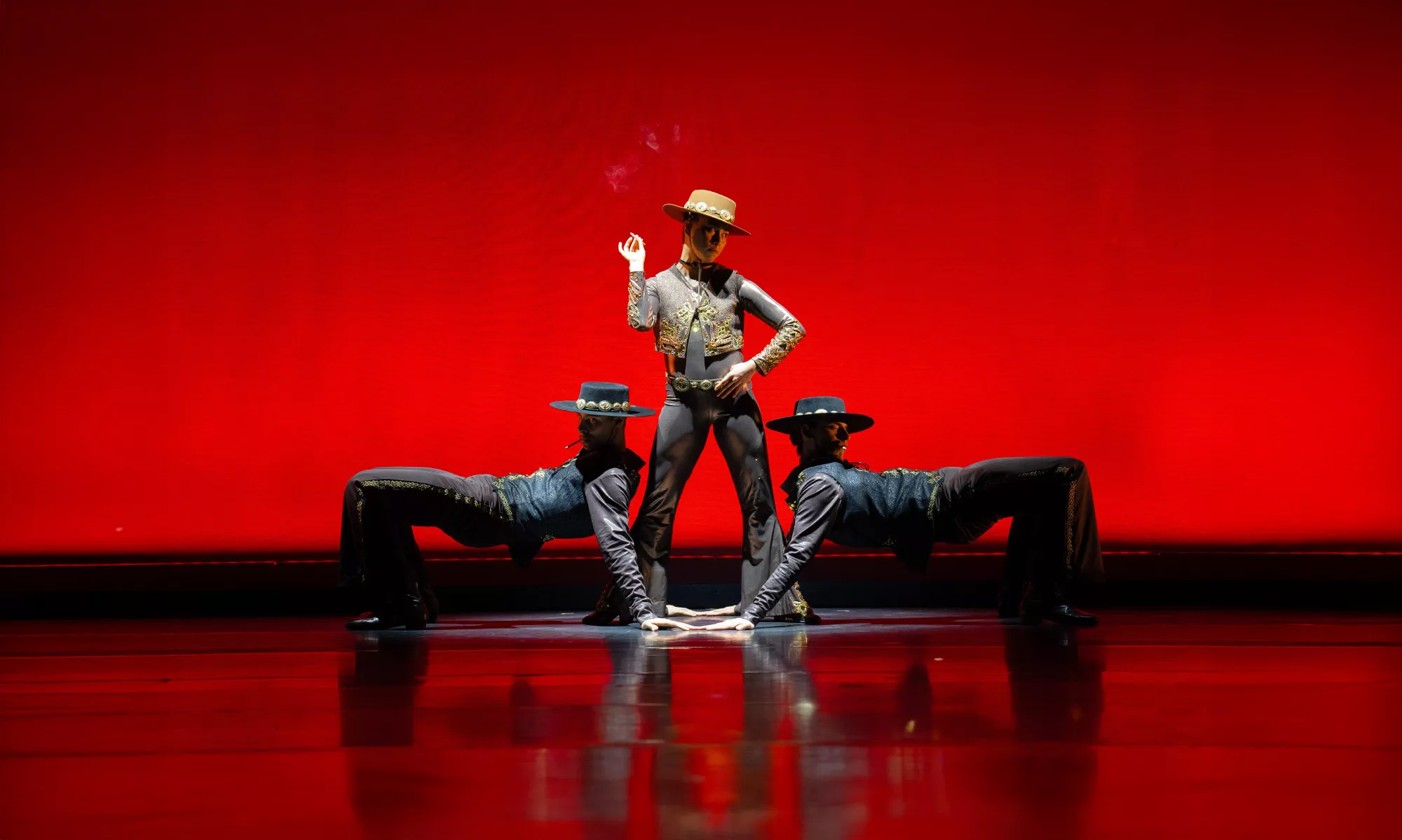 One dancer in a hat stands confidently in front of a deep red backdrop, flanked by 2 other dancers on either side.