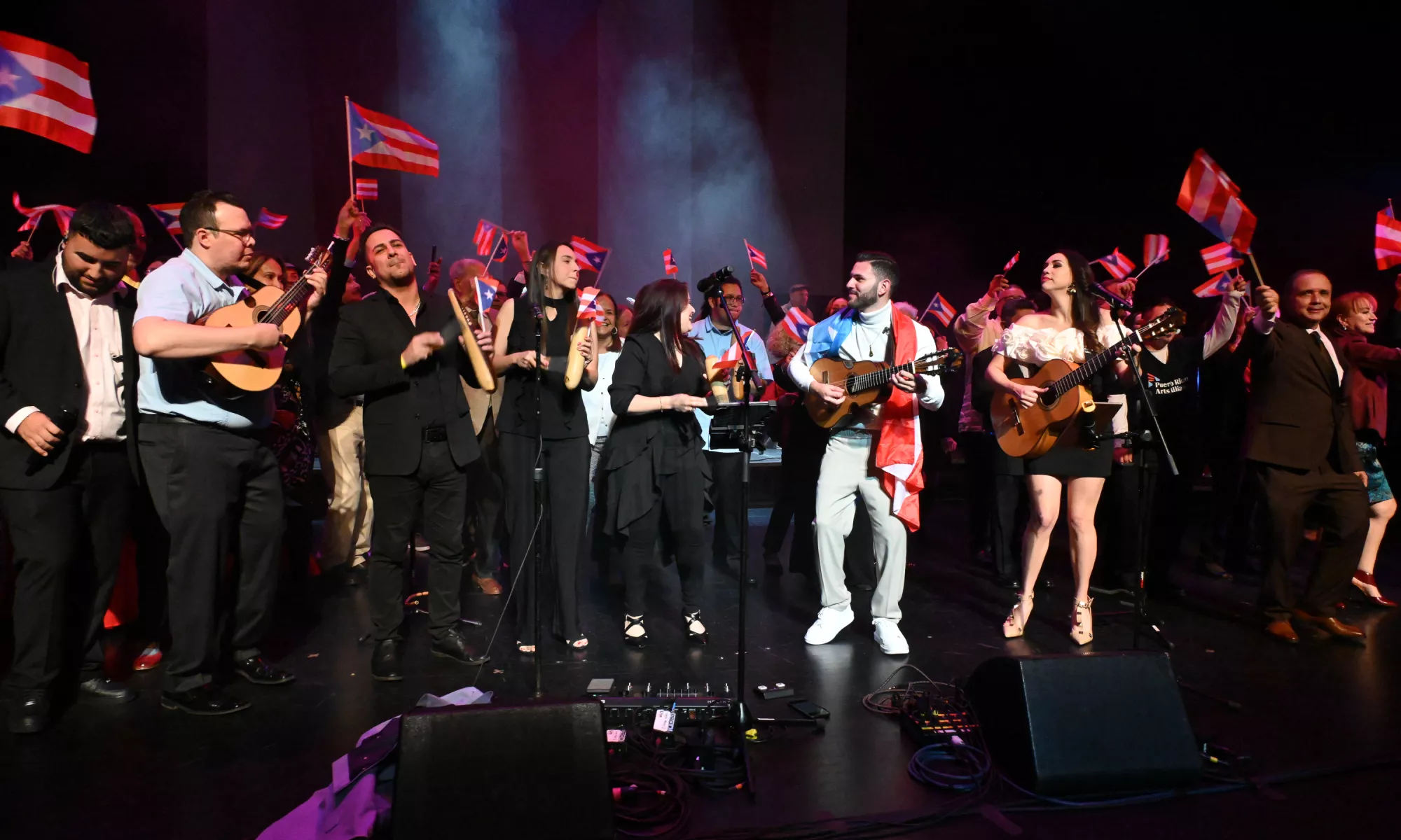 A group of people playing on stage; some play instruments while others wave or wear the Puerto Rican flag.