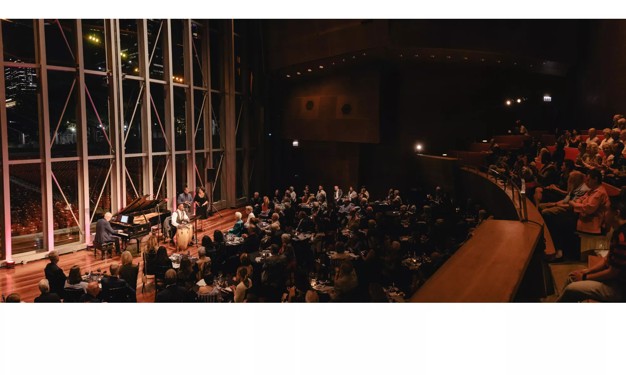 An audience seated on risers and tables watching a performance on the Pritzker stage with the Millennium Park and city skyline at night as the backdrop.