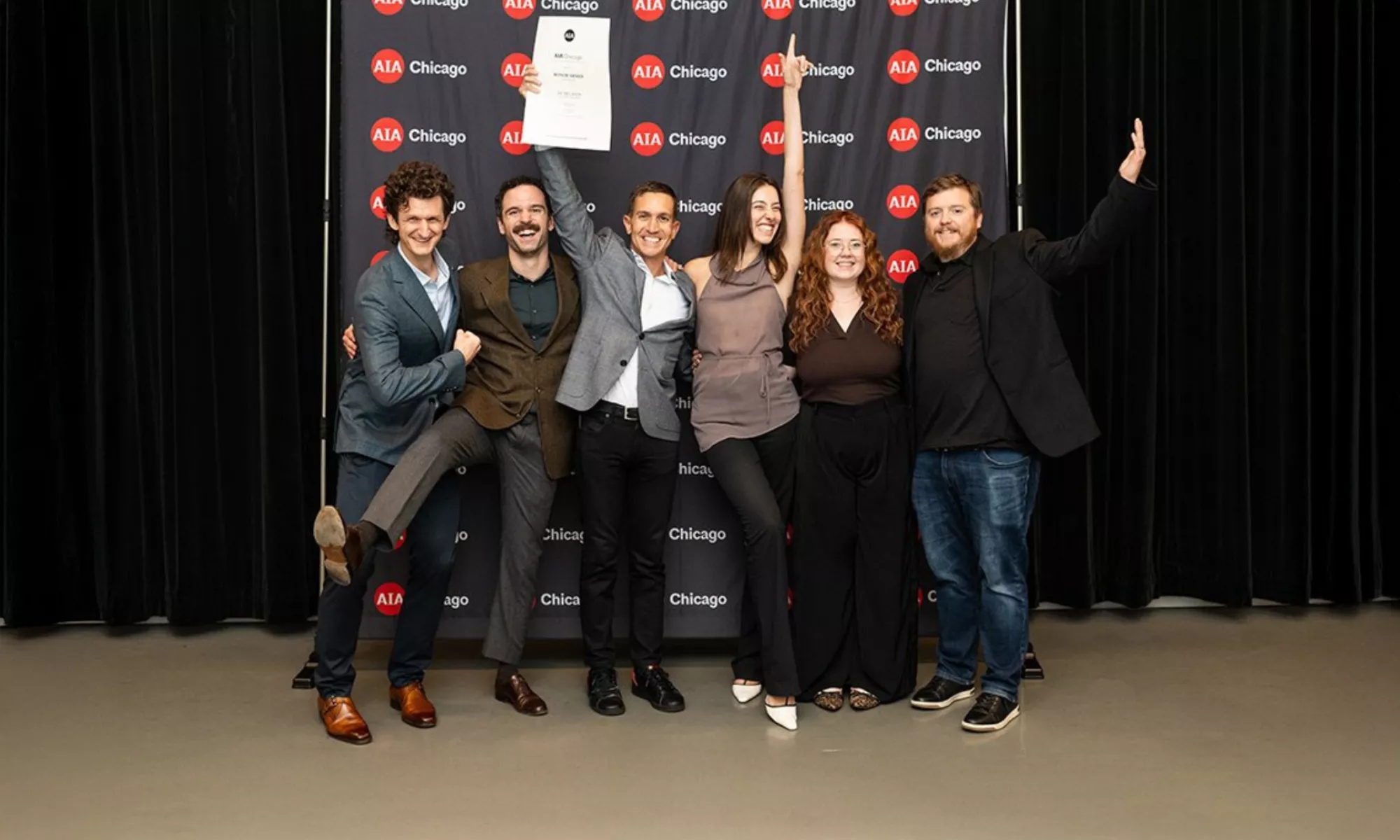 Six people celebrating their award at AIA Chicago's Designight, posing together in front of a backdrop with repeated AIA Chicago logos. Two people in the center are holding up certificates or awards while the group strikes jubilant poses with raised arms and big smiles."