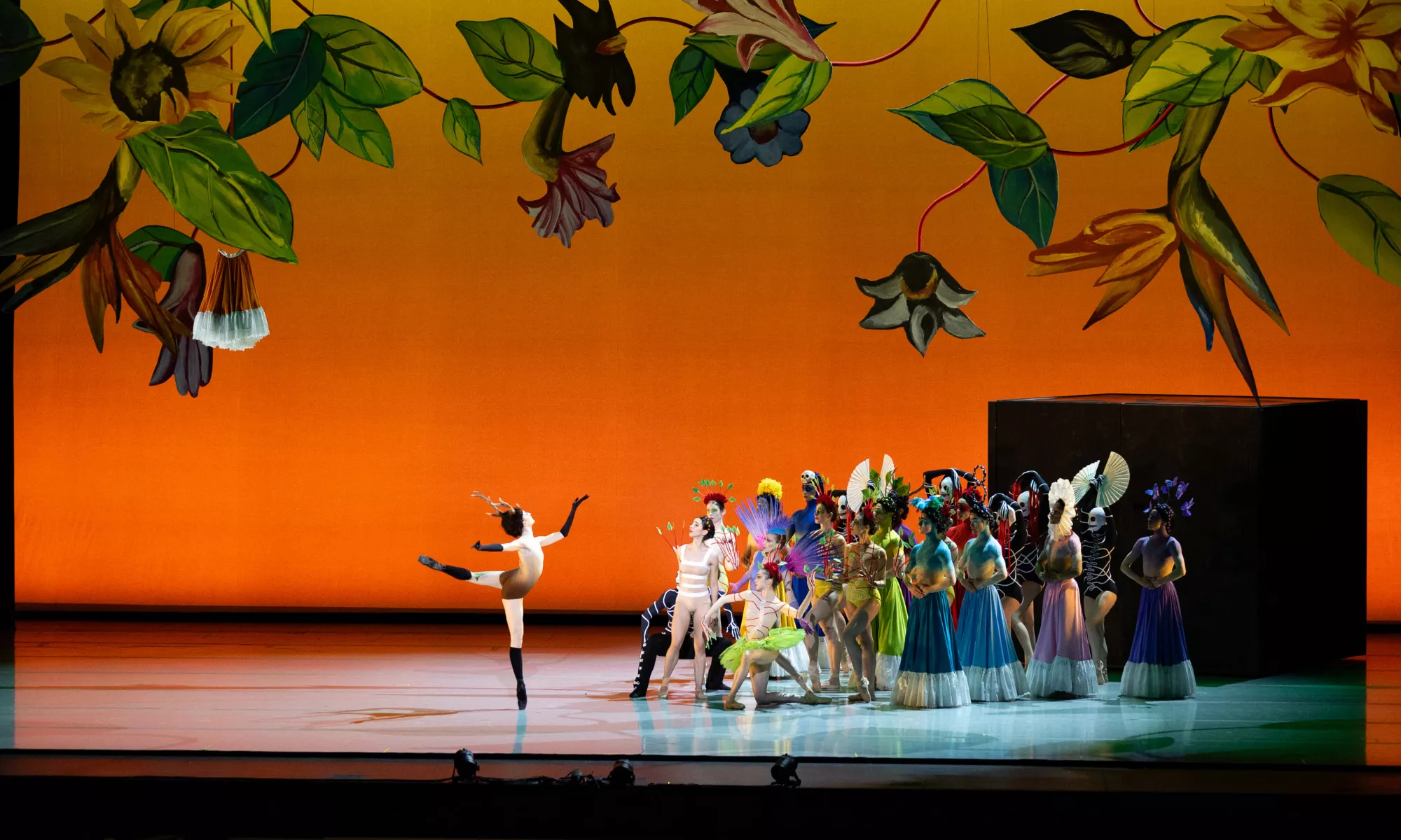 An ensemble of ballet dancers wearing eccentric costumes. A vine of leaves and flowers align the stage.