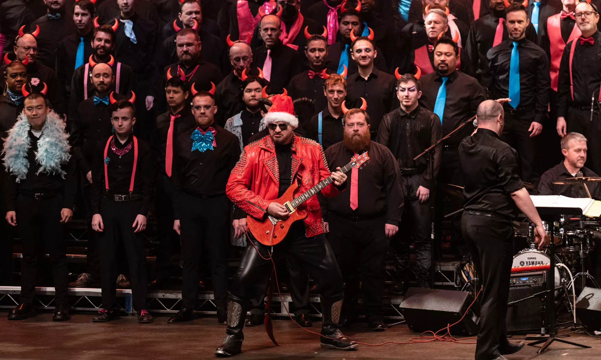 A man in a red leather jacket and Santa Claus hat plays the guitar and a choir stands behind him.