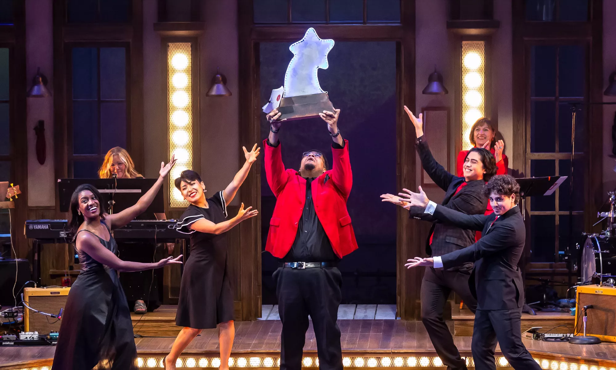 A man wearing a red blazer holds up a large star award astonished. Several others motion to him in excitment.