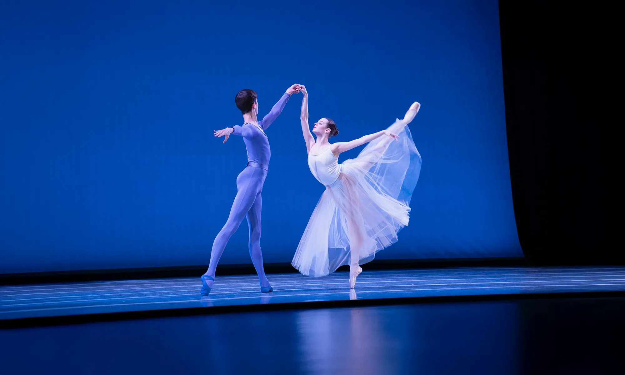 Two ballet dancers perform on stage. The ballerina holds a high arabesque, her tulle skirt sweeping around her feet, while her partner supports her at her side.