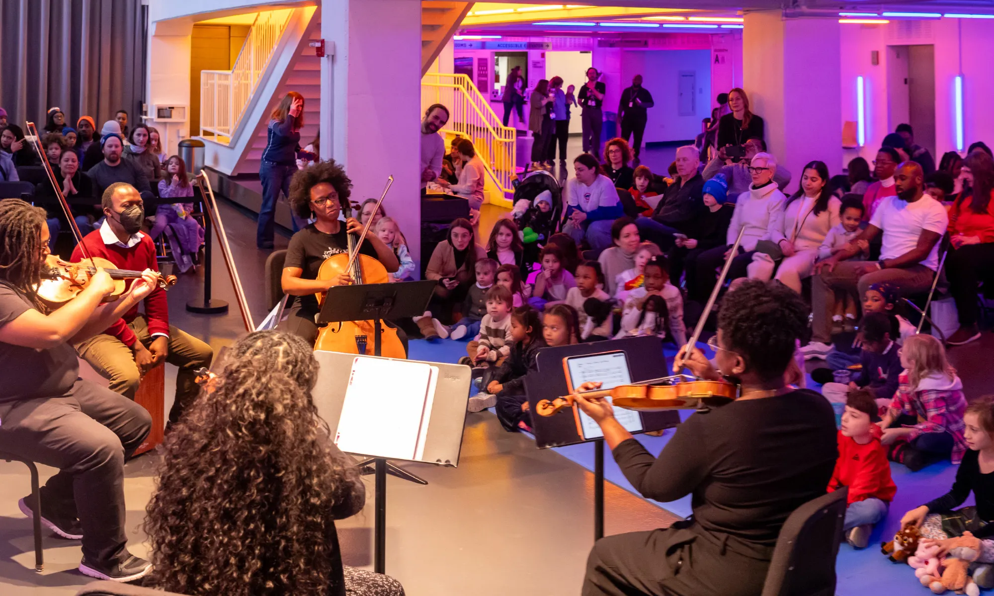 D-Composed playing in front of kids sitting on floor mats and an audience seated in the Harris Theater lobby.
