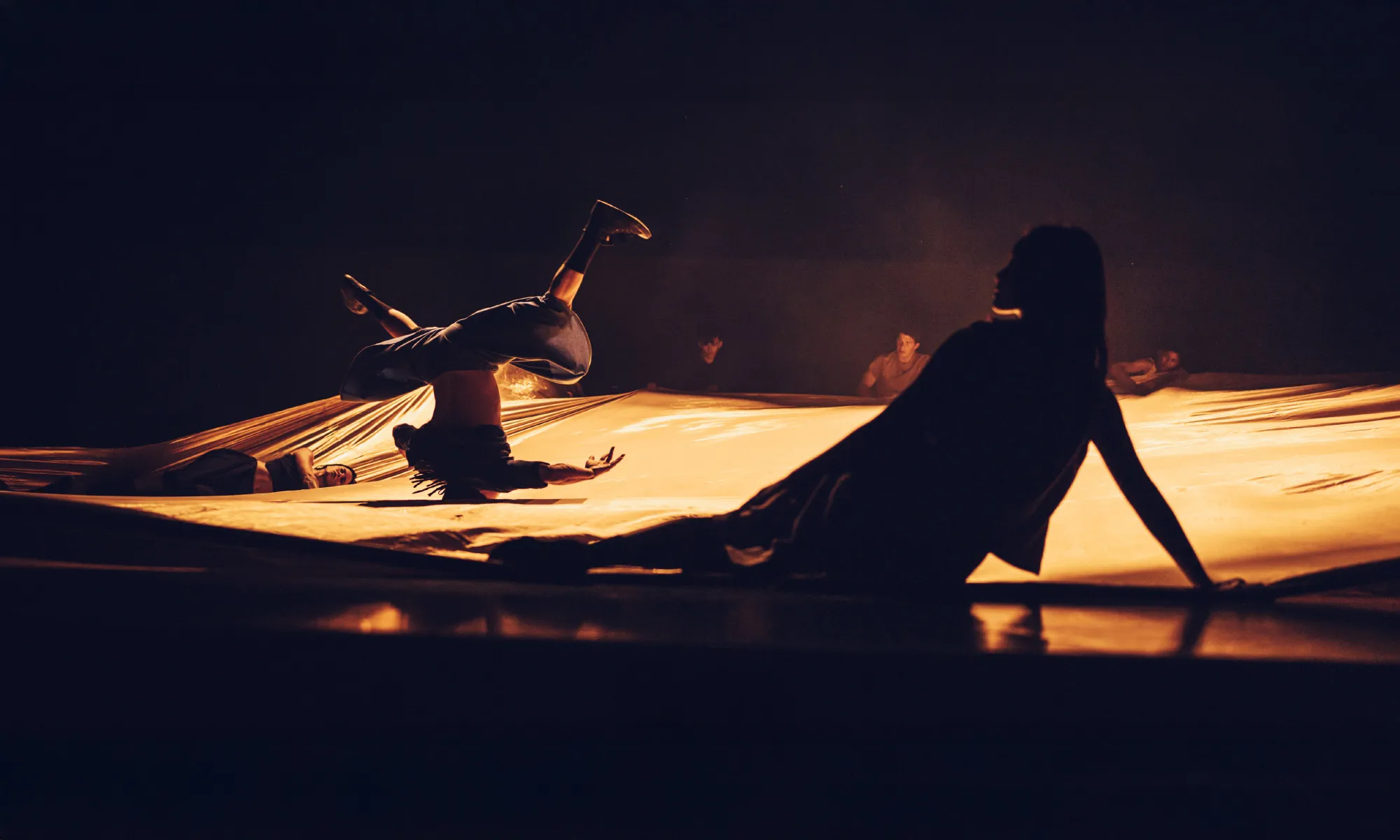 A dancer resting on the stage floor while another dancer spins on their head on top of a large drape.