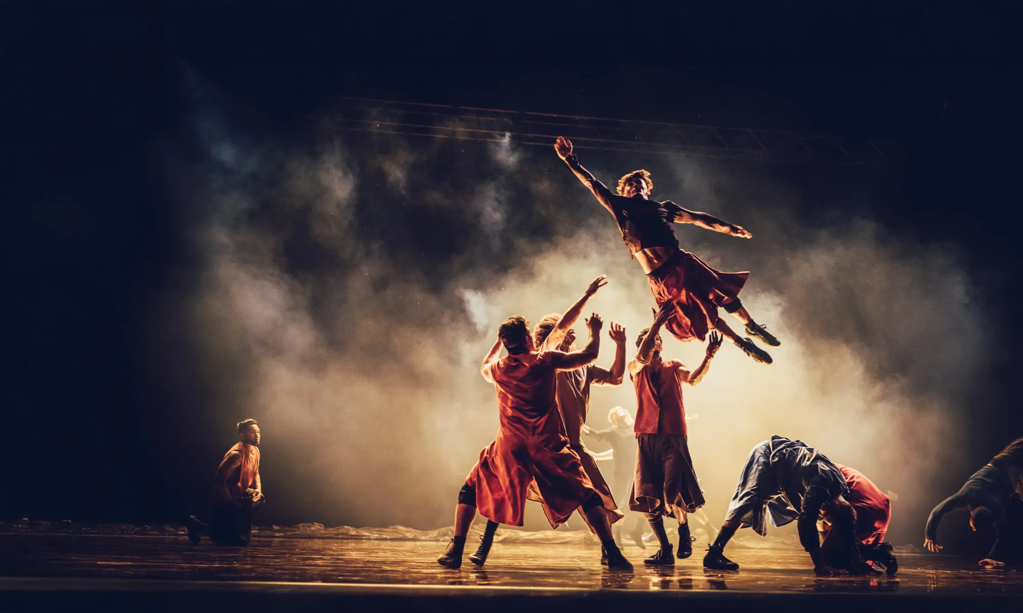 A dancer jumping in the air and four other dancers catching him. Fog covers them all.