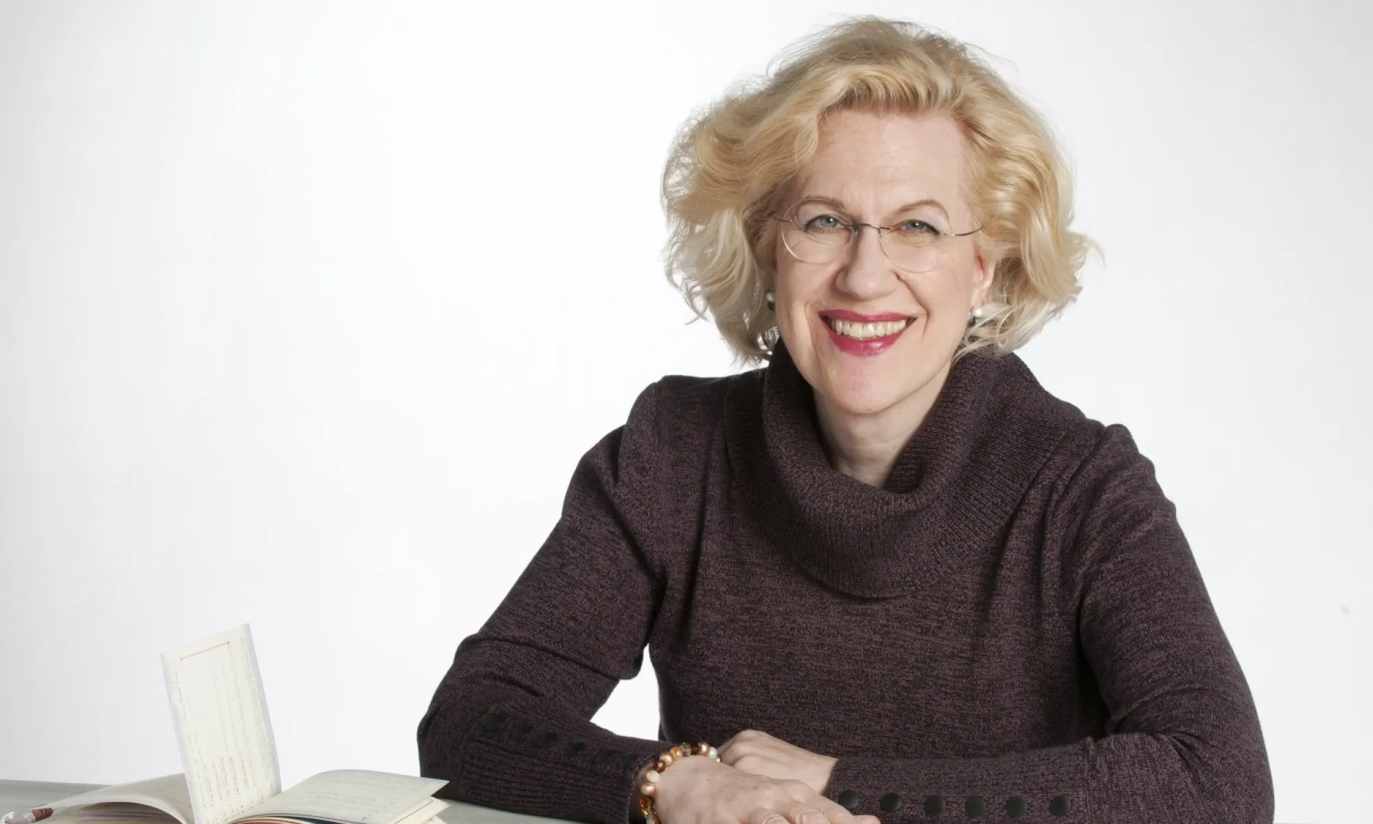 A woman smiling. She sits at a desk with a book of music sheet open.