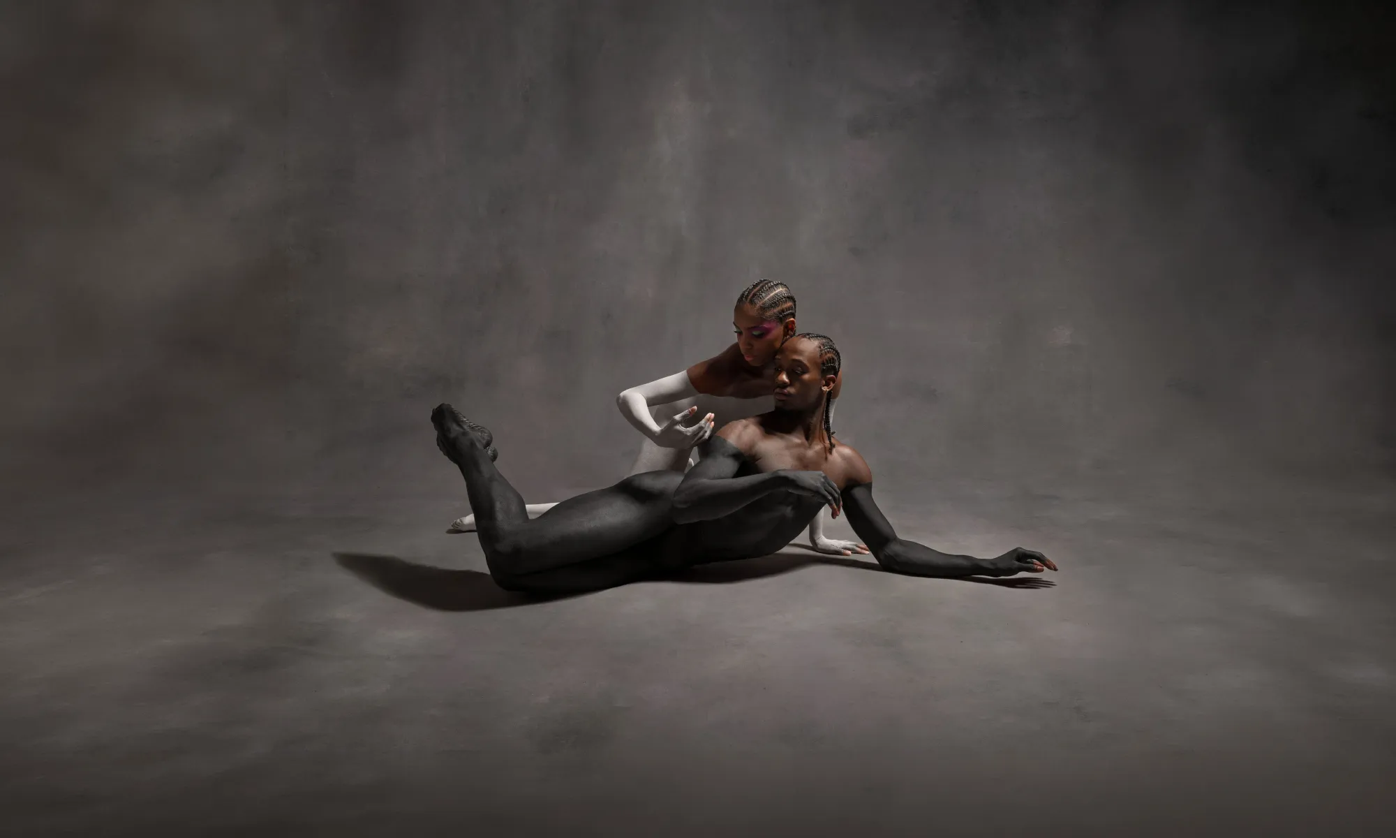 Two dancers lay on a gray backdrop. One dancer is covered in black paint up to their shoulders. The second dancer is covered in white paint.