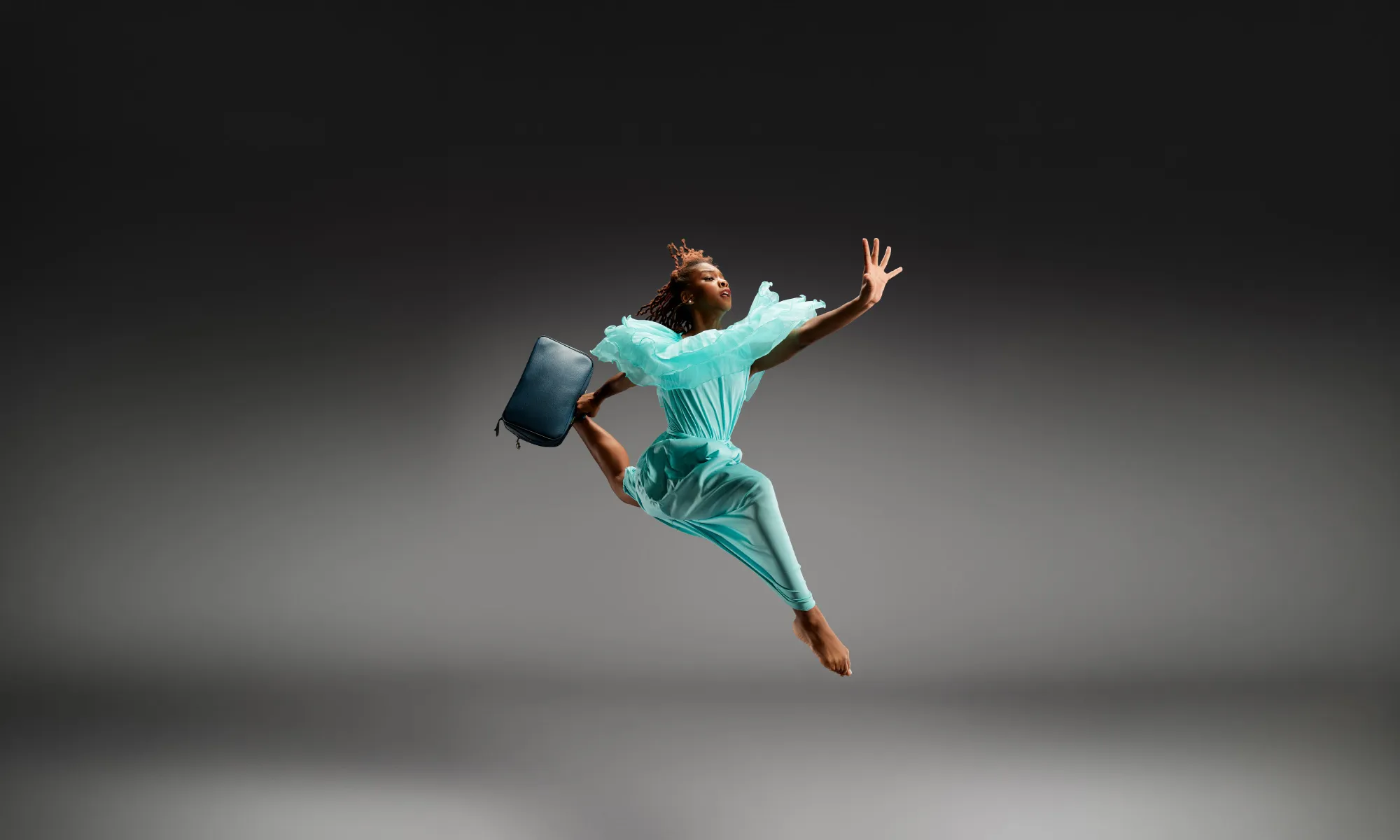 A young dancer jumping in the air wearing a blue dress and carrying a suit case.