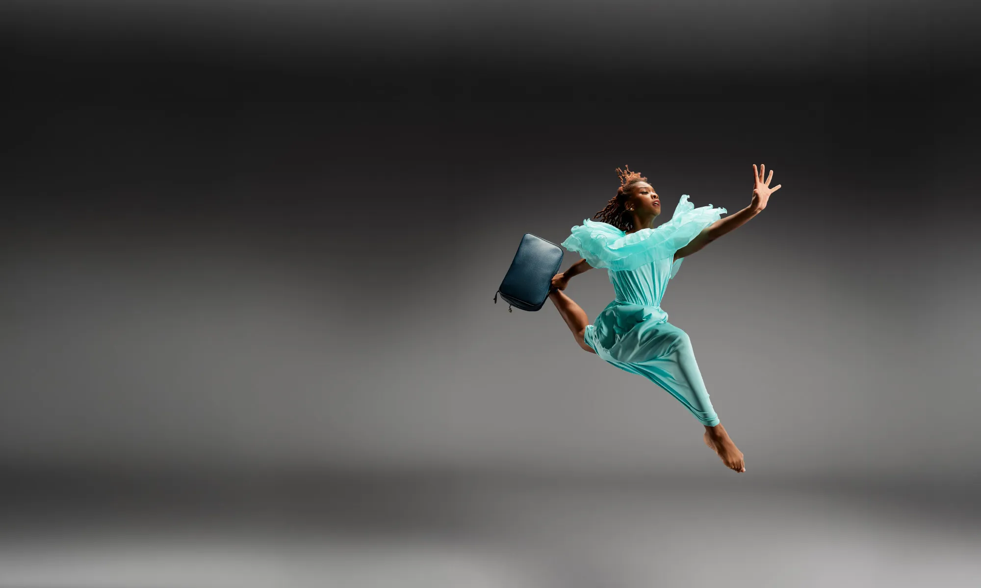 A dancer jumping in a teal dress. She carries a suit case.