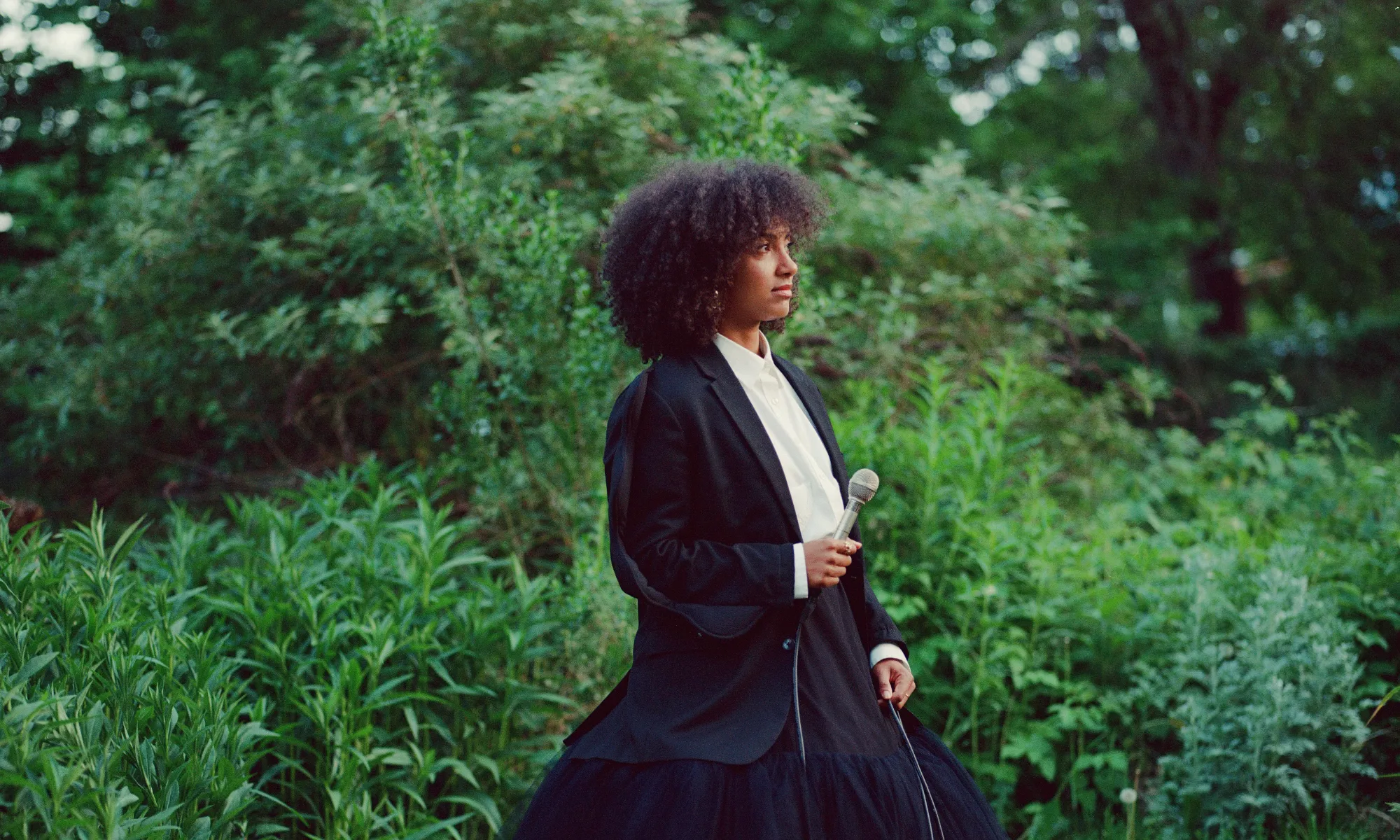 A woman in a black and white gown holding a microphone in the middle of the woods.