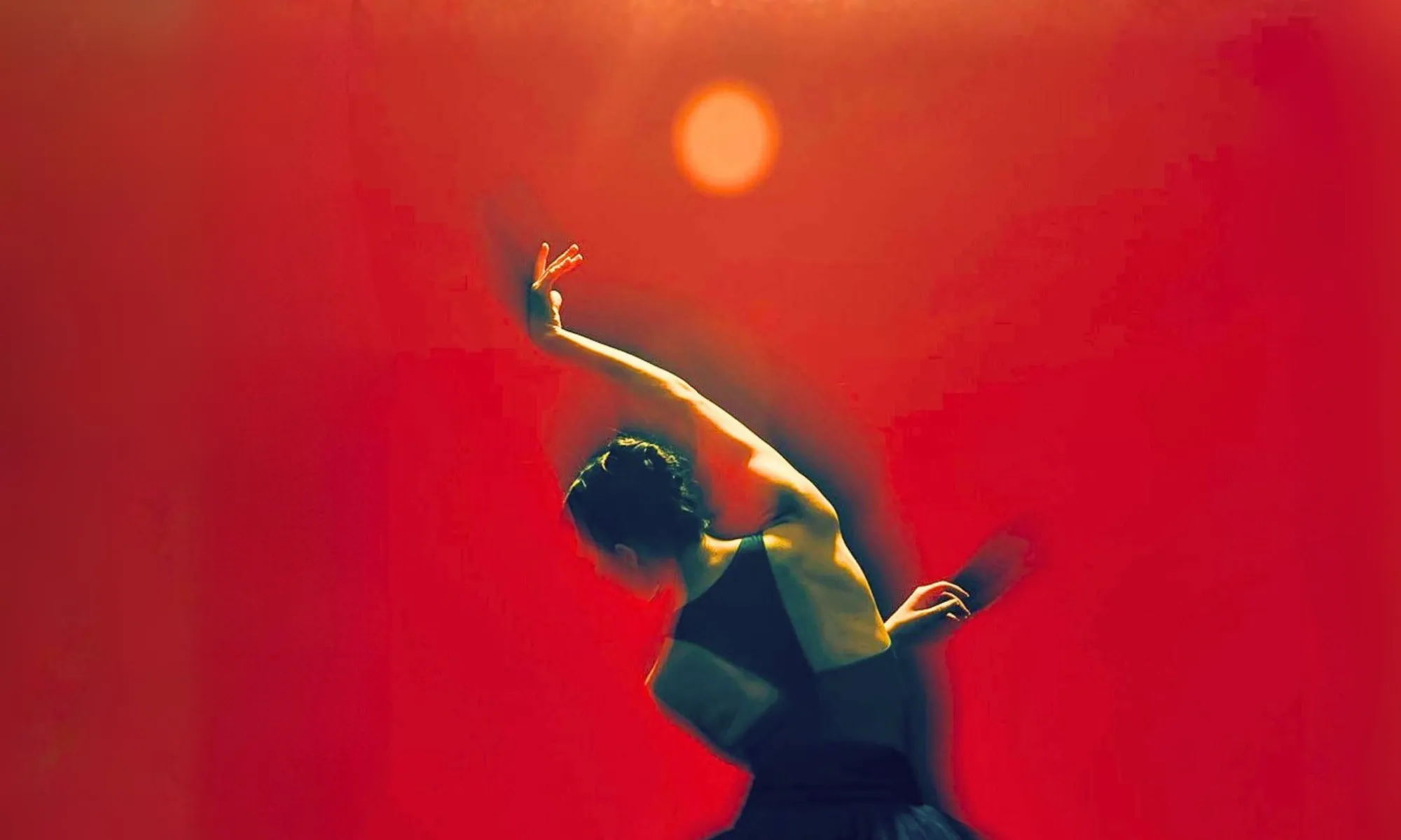A dancer in a black costume moves in a dramatic, curved pose against a vivid red-orange background. Warm light illuminates the dancer’s outstretched arm and upper body, while a glowing circular light hovers above, creating a striking, abstract composition that evokes intensity, movement, and emotion.