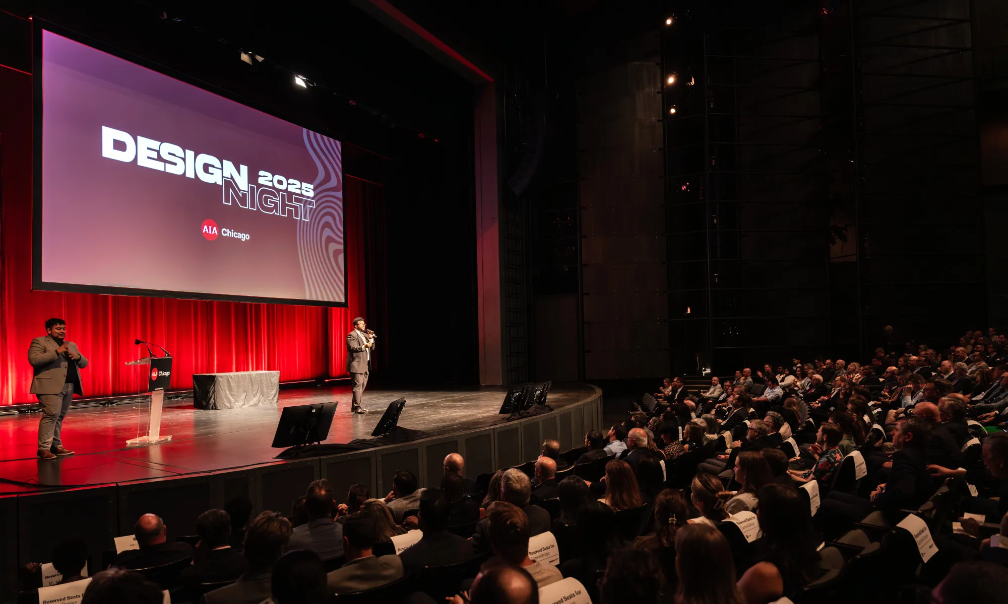 Wide shot of a large auditorium audience seated and facing a stage with a "Design 2025" screen display, a speaker standing at the podium.