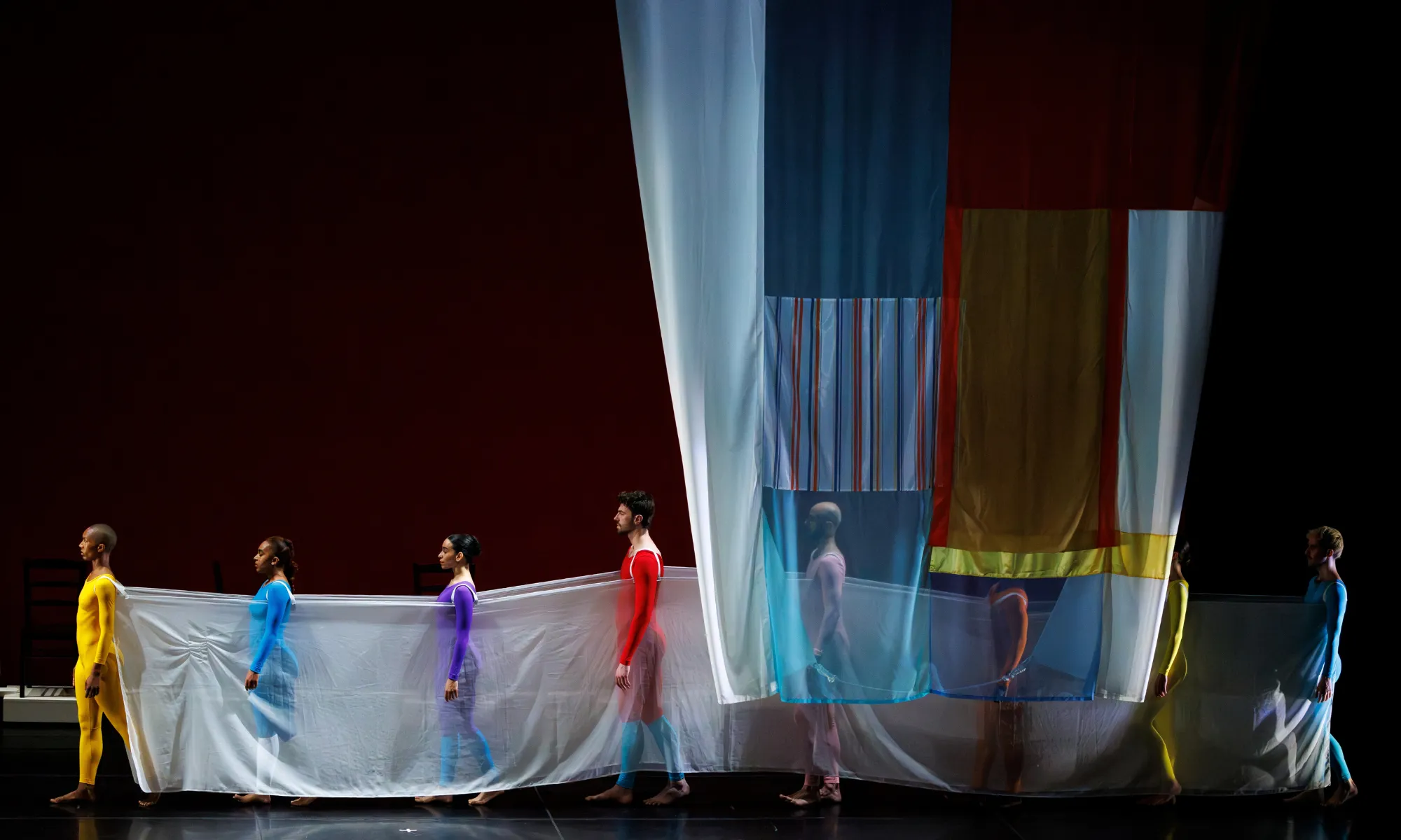 Dancers wear different color leotards while standing in a line. They hold a white cloth.
