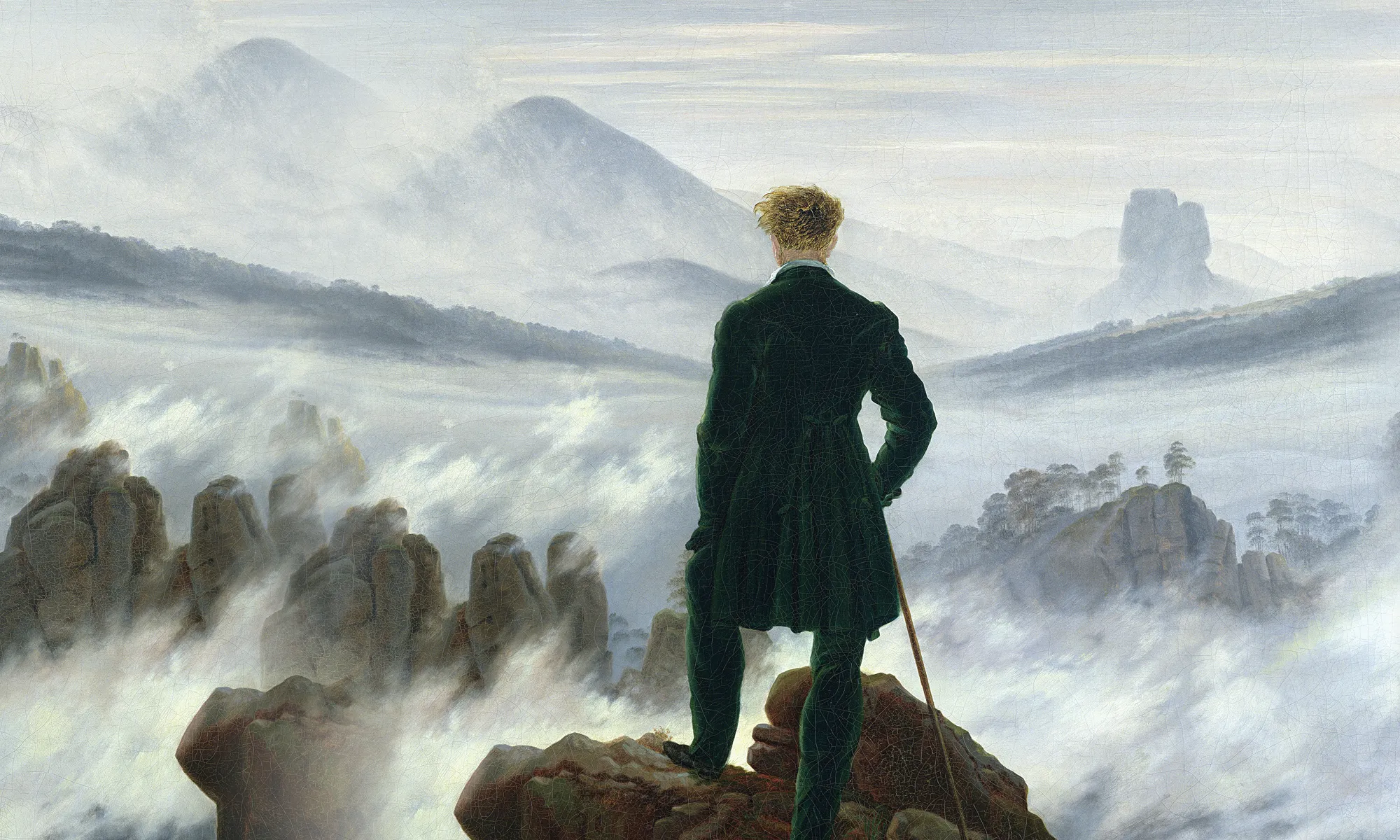 with his back to the viewer, a man stands on a rock staring out at foggy mountains