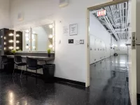 Image of Dressing Room 2. There is a vanity mirror station with chairs and a black locker to the left of the vanity. Through the open door on the right-side you can see the hallway. 