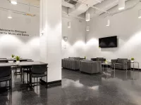 Image of an empty lounge room. On the left-side of the room, four tables with chairs are placed. On the right-side of the room four stripped couch chairs with side tables are placed. A tv hangs the wall of the right-side of the room.