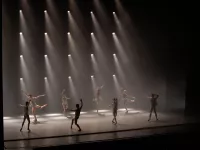 Image of eight dancers on stage wearing a plain white shirt and briefs. Several spotlights light the stage while the rest of the stage is dark. 