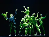 A group of dancers in bright green full length leotards. Once climbs a top of another. 