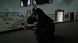 A woman with dark hair and clothing crouches with her palm at her forehead, with a large room filled with debris in the background