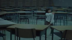 A young girl with dark hair and a white sweater dances, surrounded by desks in a classroom