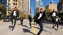 Seven Giordano Dance dancers perform in the middle of a Chicago street, tall buildings and blue sky in the background. Dancers wearing black suits with white undershirts, all caught jumping in midair.