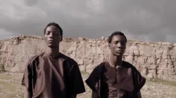 Two dancers wearing dark brown tops stand outside on rocks and grass. They are in a row facing the camera with eyes closed and  faces still. In the background, a wall of pale rock can be seen, below a sky of rolling gray clouds. 