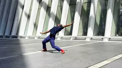 Dancer wearing blue jeans, blue button-up shirt, and orange tennis shoes stands in an empty, sun-filled parking lot. Dancer's legs are spread wide as he leans dramatically backwards, arms extended to his sides and head angled away from the camera. 