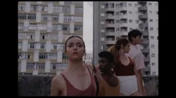 Four dancers stand close together in a diagonal row on a rooftop, tall buildings flanked behind them in a gray sky. Female dancers wear solid red or gold tops, and one male dancer wears a cream button down. One dancer looks at the camera while the others look  to the sides. 
