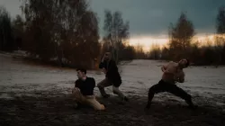 Three men in black and tan clothes dance on rocky terrain, with cloudy sky and low light on the horizon