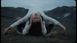 Two dancers in white shirts lie on their backs on dark green earth, one with arms extended over the other