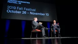 Two men, one White and one Black, sit in chairs on a dark stage, against a backdrop that reads Chicago Humanities Festival - 2019 Fall Festival
