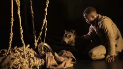 A Black man in military uniform sits with a mask and fabrics on a dark floor