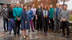 13 musicians in casual attire pose in front of trees with autumn colors