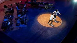 Two dancer dressed in white long sleeved shirts and black skinny jeans with white sneakers raise one arm forward, facing the audience