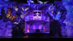 An image from the opera Florencia en el Amazonas: A woman stands with open arms at the front of a ship. She is surrounded by giant butterflies and illuminated in a purple light. 