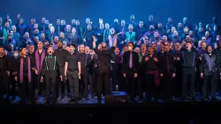 Picture of the chorus performing together onstage; members are dressed in all blacks with pops of green and purple color. Their mouths are open in song. The conductor stands at the front and center, back to the audience, arms stretched upward.