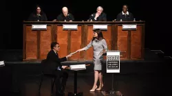 Image of participants on a stage, one is handing a file to a seated participant, with four judges in the background observing.