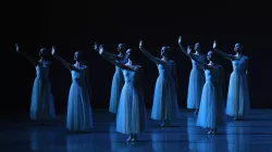 Several dancers dressed in white dresses lift their arms to stage right.