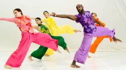 Five dancers in brightly colored, floral-appliqué costumes reach back with their arm while lunging forward with their leg. 