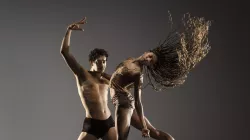 Male dancer wears black shorts, holding female dancer wearing a brown crop top and brown shorts