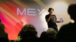 A man on stage holding a piece of paper speaks in front of crowd. The background is a multi-colored, primarily red image with the logo that reads "MEX talks".