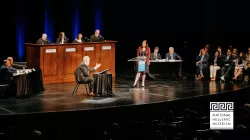 On a stage, a courtroom like setup takes place. 