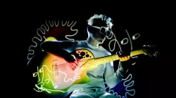 A person plays the guitar. A colorful illustration is projected on them and the guitar. 