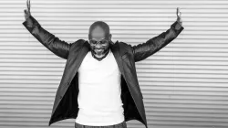 A black and white image of Marc Bamuthi Joseph while he lowers his face and extends his arms outward.