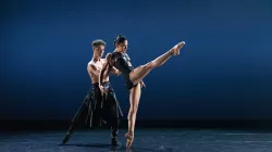 Two dancers performing. The ballerina has her leg extended while slightly leaning back onto the arms of the male dancer.