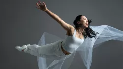 A dancer midair spread her hands open. She wears white pants and shit holding a white sheer piece of fabric. 